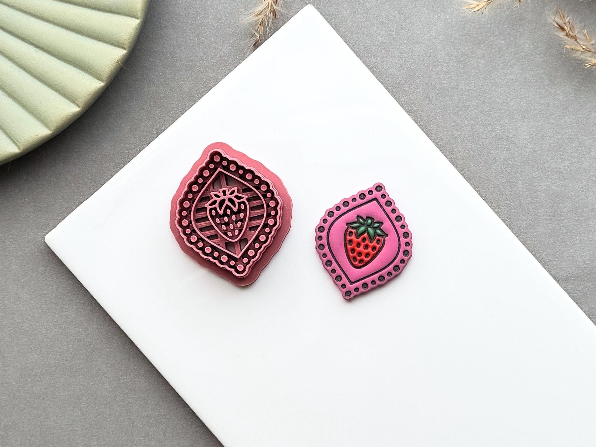 Strawberry frame polymer clay cutter displayed beside a matching finished polymer clay embellishment featuring a strawberry inside a decorative beaded frame.