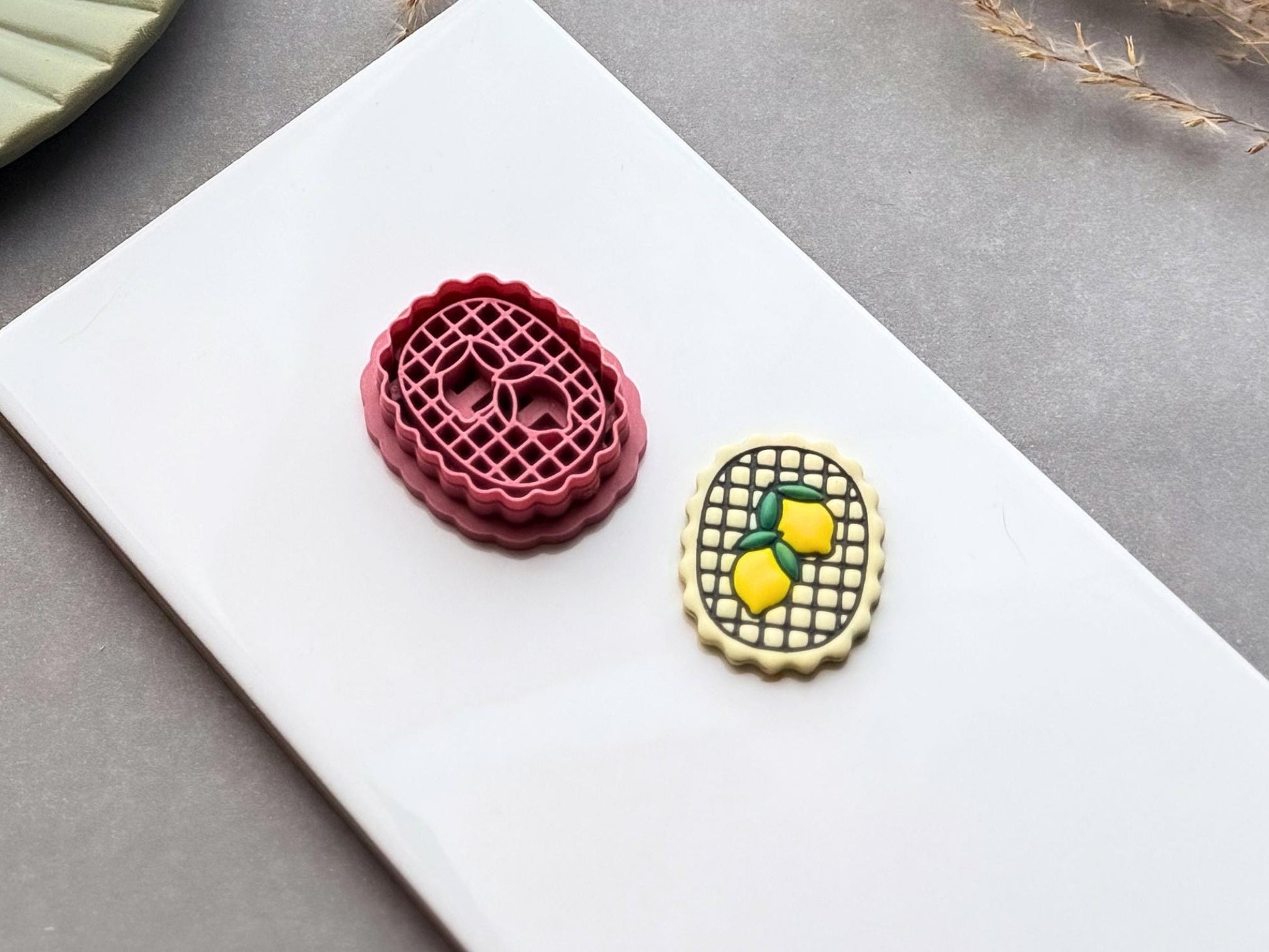Side view of a retro style scalloped oval clay cutter with two lemons and a checkered background. Next to it is a finished polymer clay earring. Both are on a white baking tile.
