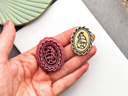 Burgundy polymer clay cutter with an ornate oval cameo frame and vintage sailing ship design, held in a hand beside the finished polymer clay cameo for size comparison.