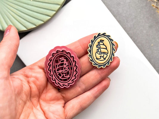 Burgundy polymer clay cutter with an ornate oval cameo frame and vintage sailing ship design, held in a hand beside the finished polymer clay cameo for size comparison.