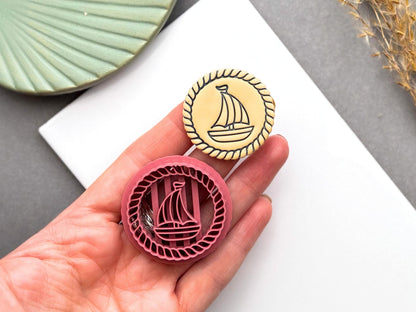Round sailing boat polymer clay cutter held in a hand for scale beside the finished clay embellishment, featuring a rope border and detailed sailboat design for nautical themed creations.