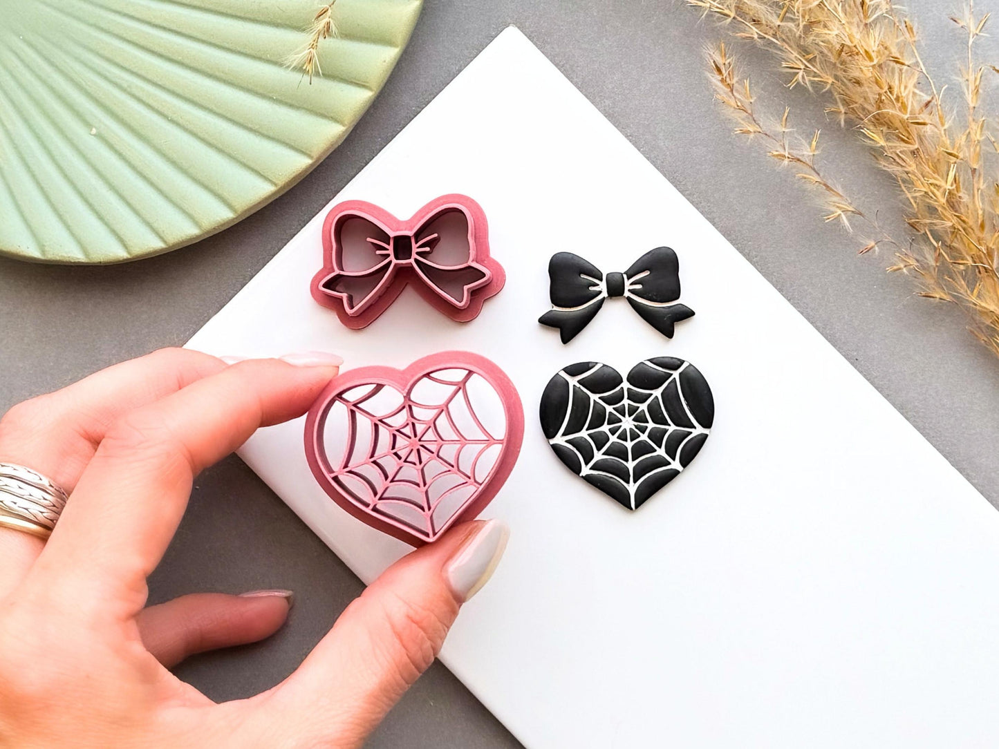 Spider web heart and bow polymer clay cutters shown alongside finished polymer clay samples, with the heart cutter held in a hand to demonstrate scale, for creating gothic jewellery, Halloween charms and spooky craft projects.