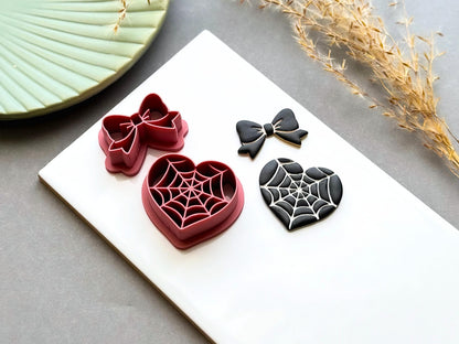 Angled side view of spider web heart and bow polymer clay cutters displayed alongside finished polymer clay samples, highlighting the layered construction and intricate spider web design for creating gothic jewellery, Halloween charms and spooky craft projects.