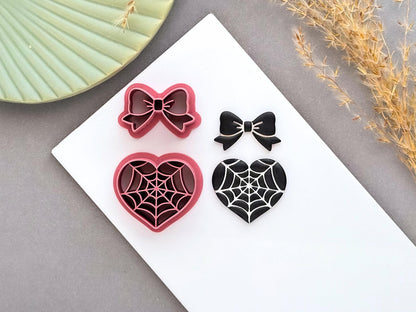 Spider web heart and bow polymer clay cutters displayed alongside finished polymer clay samples, featuring gothic spider web designs for creating Halloween jewellery, spooky charms and alternative craft projects.