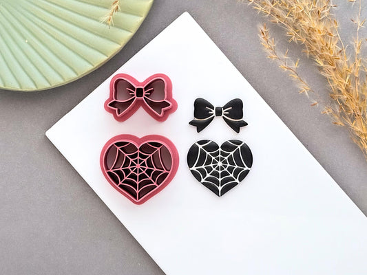 Spider web heart and bow polymer clay cutters displayed alongside finished polymer clay samples, featuring gothic spider web designs for creating Halloween jewellery, spooky charms and alternative craft projects.