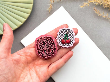 Cherry polymer clay cutter displayed beside a matching finished polymer clay embellishment featuring two cherries on a decorative lattice background within a scalloped frame.
