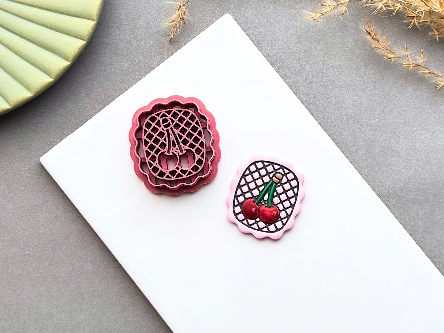 Cherry polymer clay cutter displayed beside a matching finished polymer clay embellishment featuring two cherries on a lattice background within a scalloped frame.