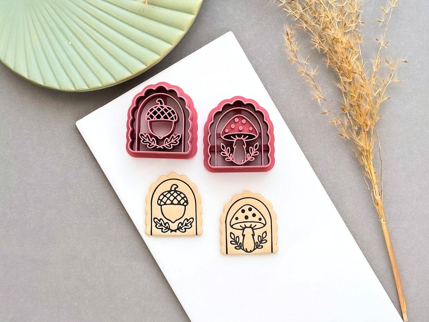 Two pink arch-shaped polymer clay cutters featuring acorn and spotted mushroom designs, shown beside matching stamped clay pieces on a white tile. Dried grasses and a green ceramic dish are visible in the background.