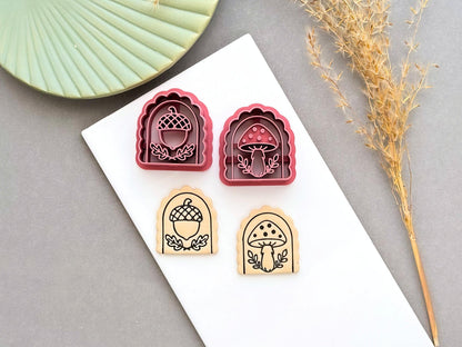 Two pink arch-shaped polymer clay cutters featuring acorn and spotted mushroom designs, shown beside matching stamped clay pieces on a white tile. Dried grasses and a green ceramic dish are visible in the background.