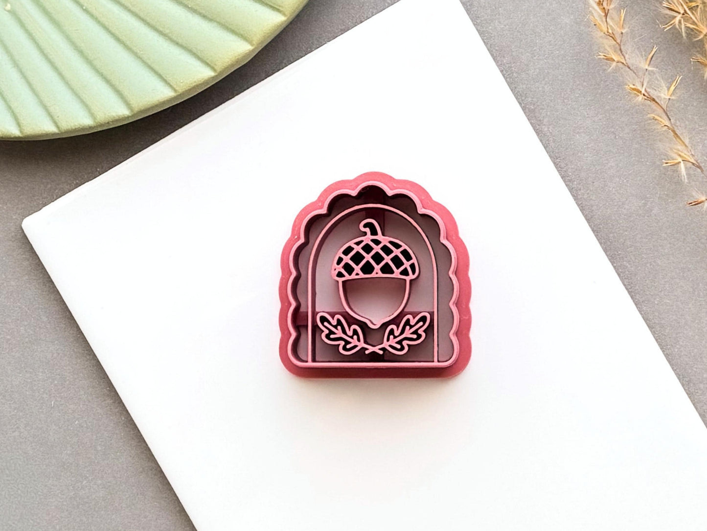 Pink arch-shaped polymer clay cutter featuring a detailed acorn design with a crosshatched cap and leafy branches, shown on a white tile.