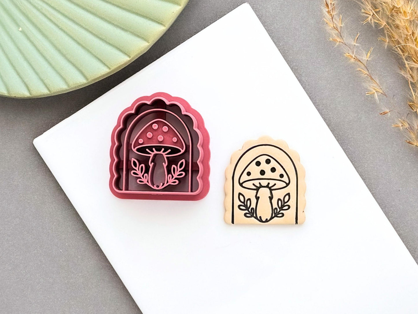 Pink arch-shaped polymer clay cutter featuring a detailed mushroom design, shown beside a matching stamped clay piece with a spotted mushroom and leafy details on a white tile.