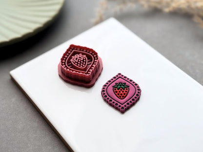 Strawberry frame polymer clay cutter displayed beside a matching finished polymer clay embellishment, with the cutter shown from a side angle to highlight the sharp cutting edge, reinforced support wall and comfortable grip handle.