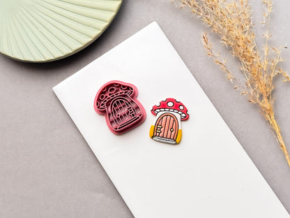 Mushroom cottage polymer clay cutter and finished clay piece, showing a red spotted mushroom with a tiny door, on a white surface.