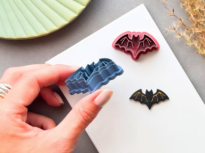 Bat polymer clay cutter held in a hand, shown from the side to highlight its layered construction and cutting edge, alongside matching finished bat clay pieces in different colours and sizes.