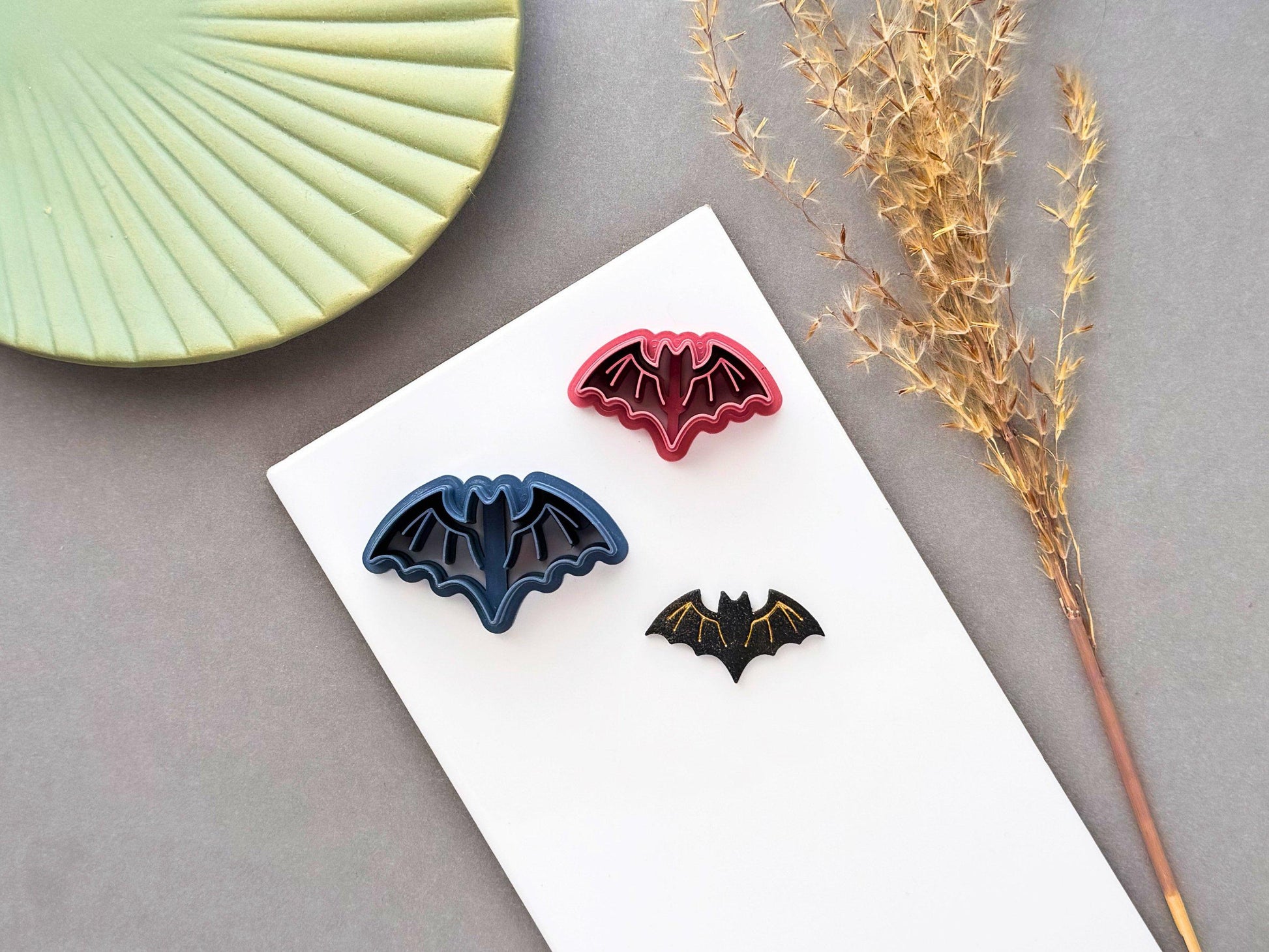 Bat polymer clay cutter displayed with matching finished clay bats in different colours and sizes, featuring a simple winged bat design, perfect for Halloween, gothic and spooky clay projects.