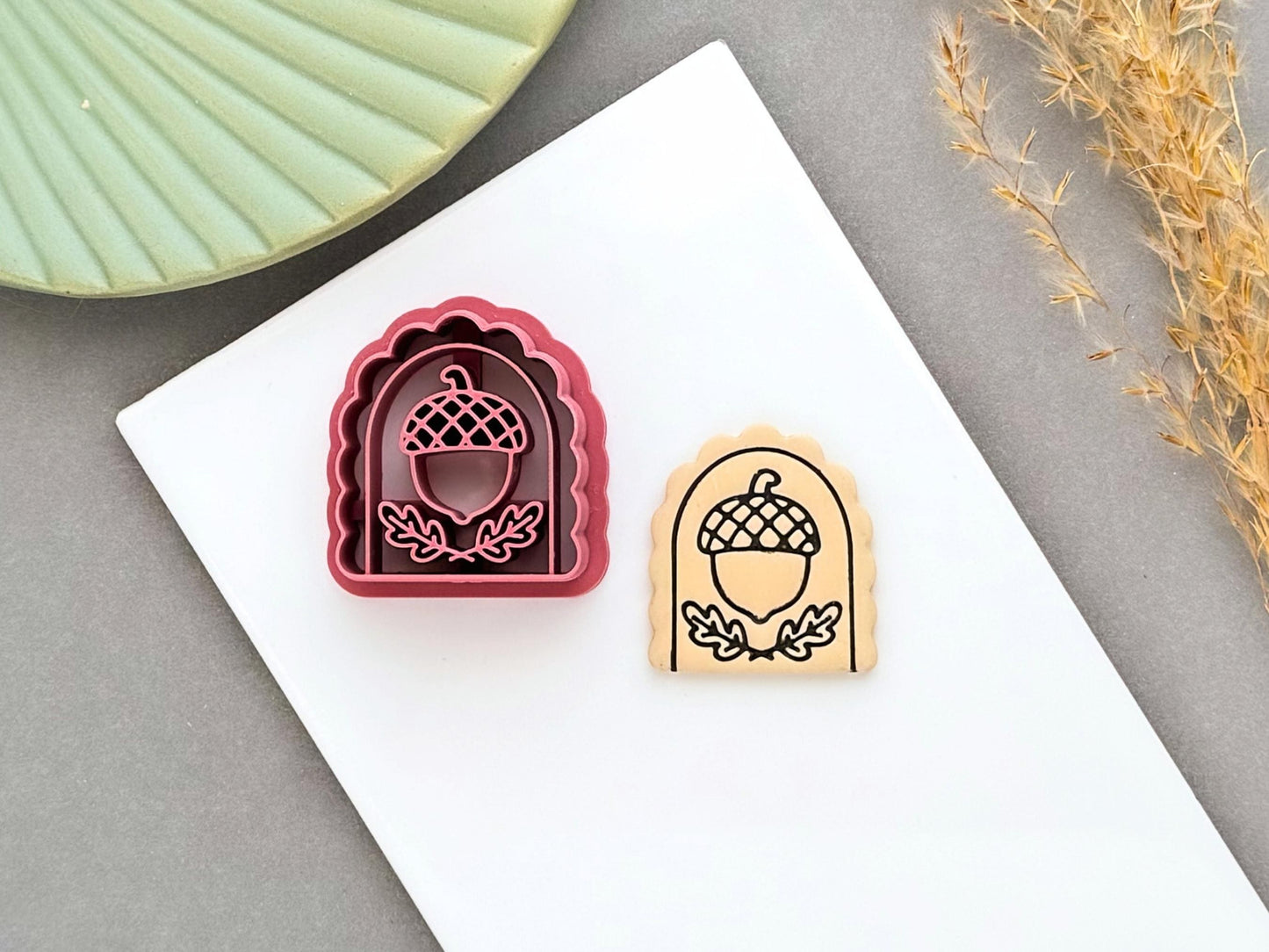 Pink arch-shaped polymer clay cutter featuring a detailed acorn design with leafy branches, shown beside a matching stamped clay piece on a white tile.