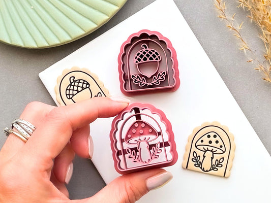 Pink arch-shaped polymer clay cutters featuring acorn and mushroom designs, shown beside matching stamped clay pieces. A hand holds the mushroom cutter to show its size and detailed cutting design.
