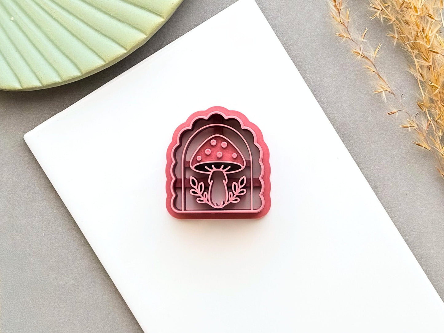 Pink arch-shaped polymer clay cutter featuring a detailed spotted mushroom design with leafy branches, shown on a white tile.