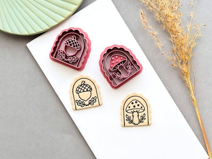 Pink arch-shaped polymer clay cutters featuring acorn and mushroom designs, shown beside matching tan clay pieces stamped with detailed acorn and mushroom motifs on a white tile.