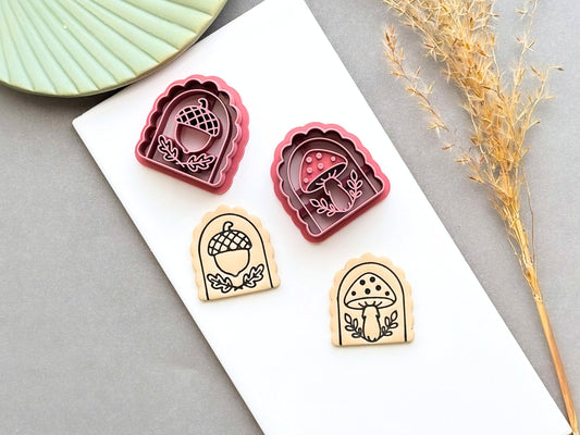 Pink arch-shaped polymer clay cutters featuring acorn and mushroom designs, shown beside matching tan clay pieces stamped with detailed acorn and mushroom motifs on a white tile.