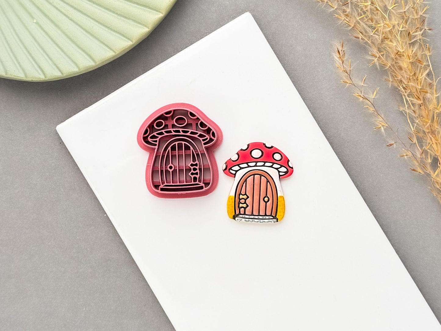 Mushroom cottage polymer clay cutter displayed beside a matching finished clay embellishment, featuring a whimsical red-and-white spotted mushroom with a tiny arched door, perfect for cottagecore, woodland, fairycore and whimsical clay projects.