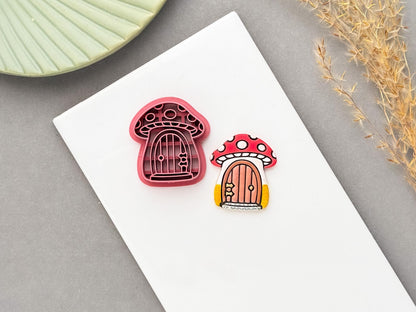Mushroom cottage polymer clay cutter displayed beside a matching finished clay embellishment, featuring a whimsical red-and-white spotted mushroom with a tiny arched door, perfect for cottagecore, woodland, fairycore and whimsical clay projects.