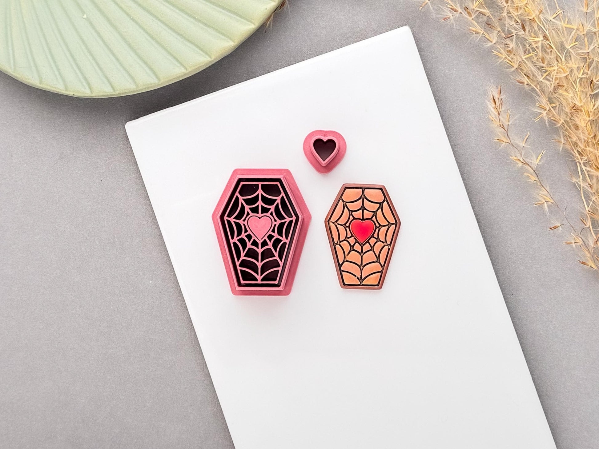 Coffin-shaped spider web polymer clay cutter with a heart centre, shown beside a matching finished clay piece and small heart cutter, on a white surface.