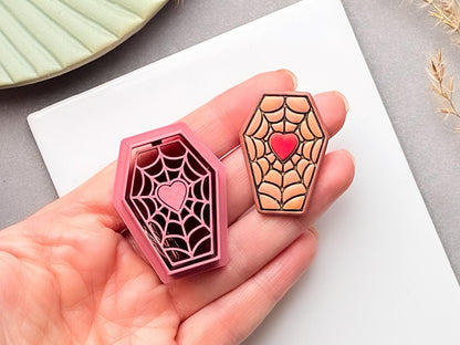 Coffin-shaped spider web polymer clay cutter with a heart centre, held in a hand beside a matching finished clay piece with a red heart.