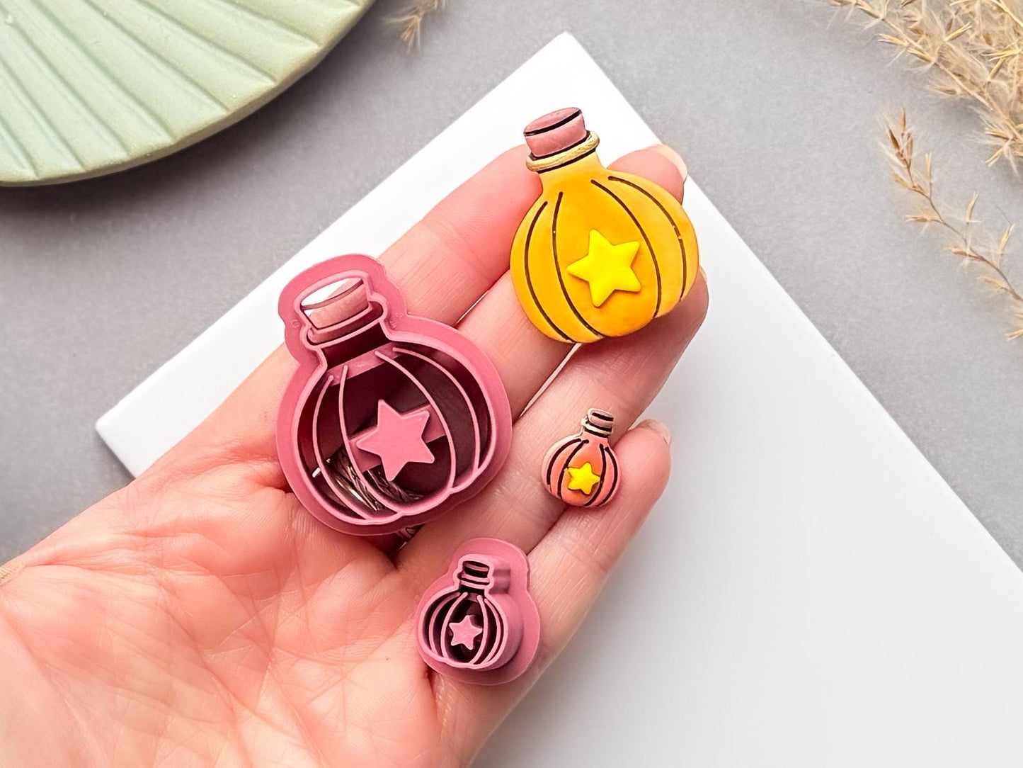 Potion bottle polymer clay cutter with star-shaped centre, held in a hand beside matching finished clay pieces in different sizes.