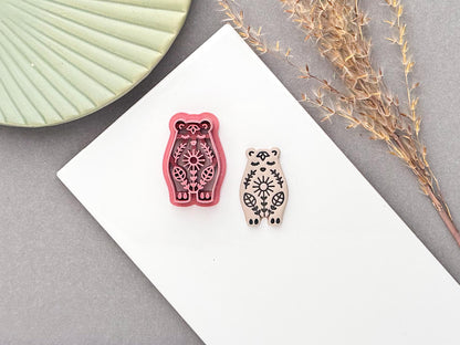 Pink bear shaped clay cutter with decorative folk style floral details next to a polymer clay sample cut out. Both are on a white baking tile.