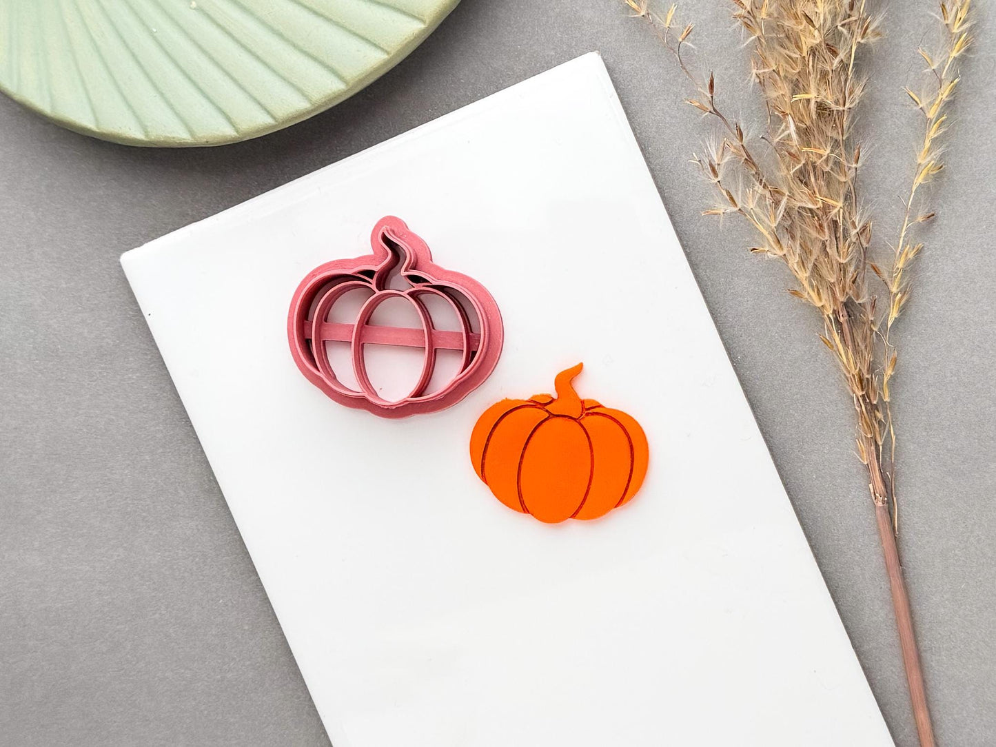 Pumpkin polymer clay cutter with a distinctive curled stem shape, shown alongside a finished orange pumpkin clay piece.