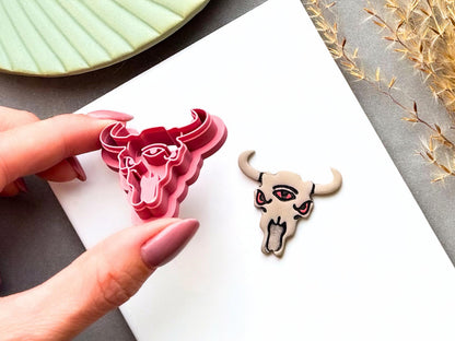 Three-eyed bull skull tilted clay cutter held in a hand, featuring curved horns, three eyes, pointed ears, facial details and a long tongue, shown beside a finished cream polymer clay bull skull with red eyes and tongue. Alternative, gothic and western-inspired design.