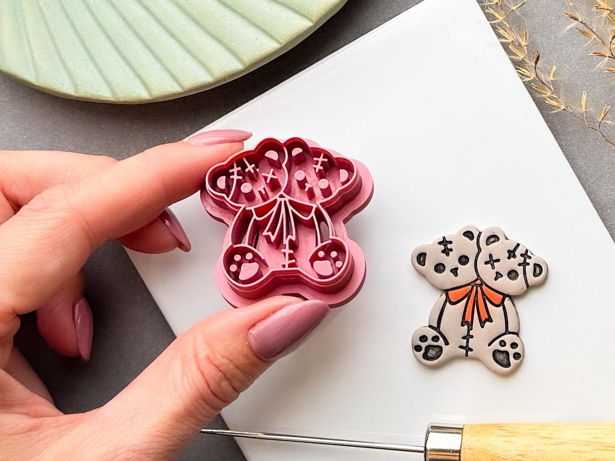 Two-headed Halloween teddy bear clay cutter held in a hand beside a finished cream polymer clay bear, featuring stitched patchwork details, cross-shaped stitches, paw pads and an orange bow tie. Spooky gothic and alternative Halloween design.