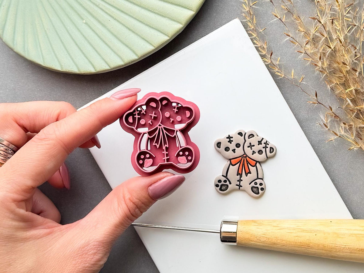 Two-headed Halloween teddy bear clay cutter held in a hand beside a finished cream polymer clay bear, featuring stitched patchwork details, cross-shaped stitches, paw details and an orange bow tie. Spooky, gothic and alternative Halloween-inspired design.