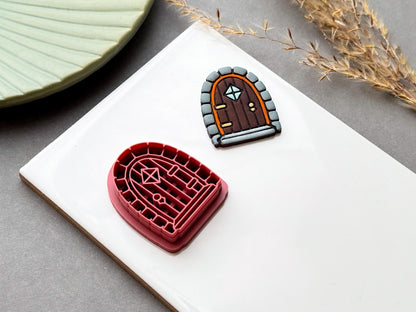Cottagecore autumn fairy door clay cutter with an arched door design featuring stone detailing, a small diamond-shaped window, and decorative door hardware, shown alongside a finished clay door.