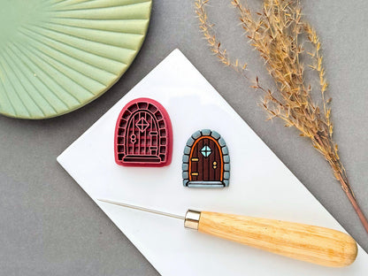 A pink plastic arch-shaped cookie cutter with an intricate arched fairy doorway design, featuring decorative panels, a central diamond-shaped window, and small geometric details. A finished brown arched door-shaped clay piece with blue-grey stone detailing is shown beside it on a white surface, with a wooden-handled needle tool in the foreground and dried foliage in the background.