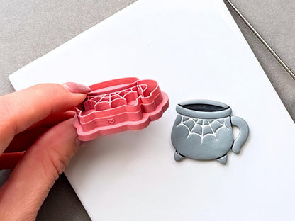 Side view of a Halloween cauldron clay cutter held in a hand, showing its layered 3D construction, spiderweb stamp details, strong support wall and raised base, alongside a finished grey polymer clay cauldron with matching web detail.