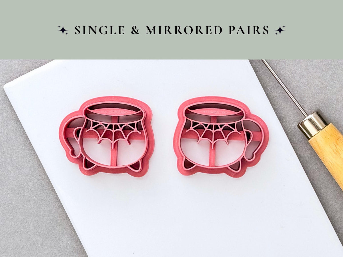 Two Halloween cauldron clay cutters shown as a mirrored pair, each featuring a spiderweb design across the front, curved handles and small feet. Pink 3D-printed cutters displayed on a white surface.