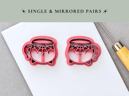 Two Halloween cauldron clay cutters shown as a mirrored pair, each featuring a spiderweb design across the front, curved handles and small feet. Pink 3D-printed cutters displayed on a white surface.