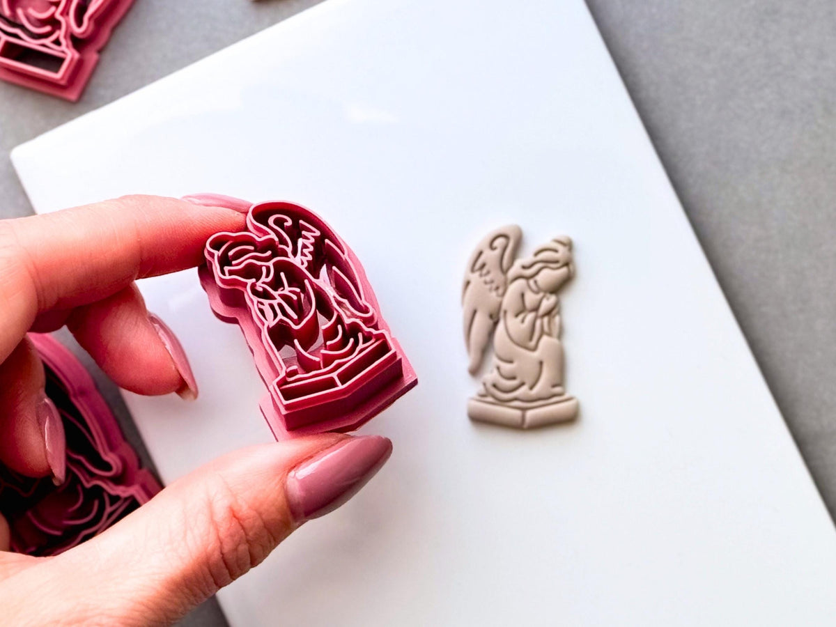 Tilted gothic angel clay cutter held in a hand, featuring a detailed winged angel statue kneeling on a pedestal, shown beside a finished cream polymer clay angel cut-out. Dark, gothic and alternative-inspired design.
