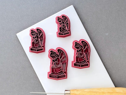 Gothic angel clay cutters displayed in four sizes, each featuring a detailed kneeling winged angel statue on a pedestal with intricate robe and feather details. Pink 3D-printed cutters arranged on a white surface.