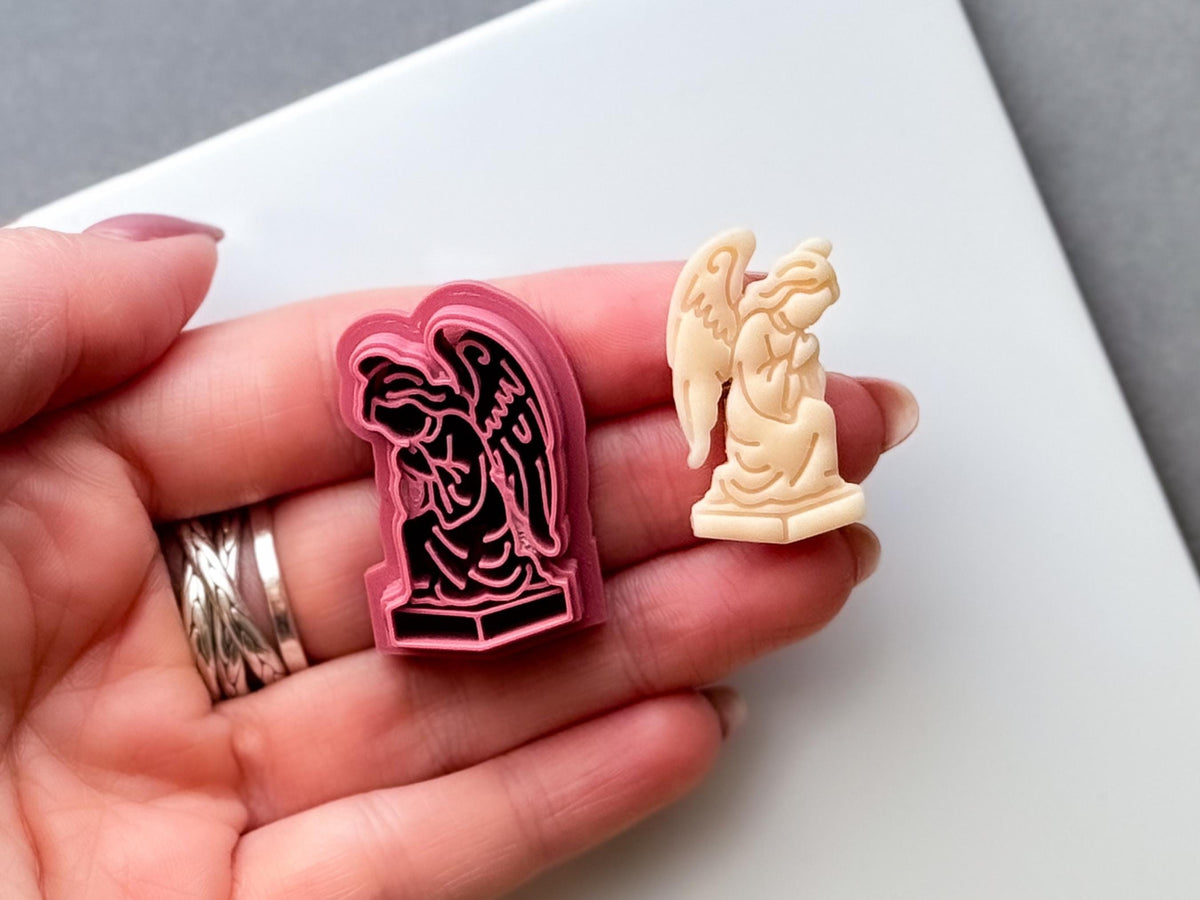Gothic angel clay cutter held in a hand, featuring a detailed winged angel statue kneeling on a pedestal, shown beside a finished cream polymer clay angel cut-out. Dark, gothic and alternative-inspired design.