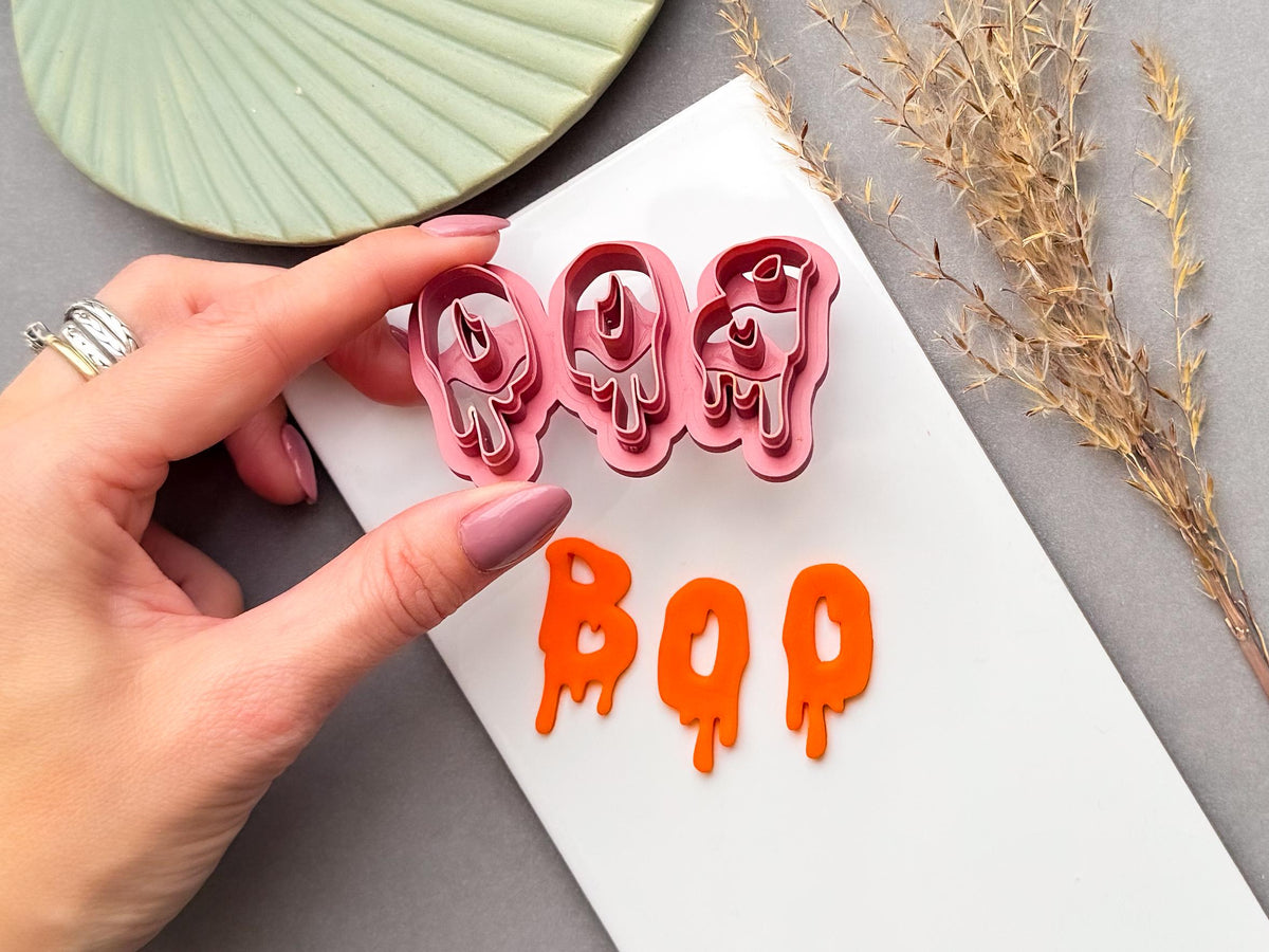 Halloween clay cutter featuring the letters “BOO” in a dripping slime-style design, held in a hand above finished orange polymer clay “BOO” letters. Spooky Halloween-inspired design for alternative jewellery and seasonal craft projects.