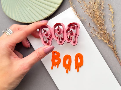 Halloween clay cutter featuring the letters “BOO” in a dripping slime-style design, held in a hand above finished orange polymer clay “BOO” letters. Spooky Halloween-inspired design for alternative jewellery and seasonal craft projects.