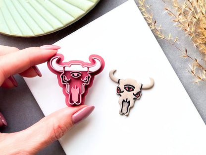 Three-eyed bull skull clay cutter held in a hand, featuring curved horns, three eyes, pointed ears, facial details and a long tongue, shown beside a finished cream polymer clay bull skull with red eyes and tongue. Alternative, gothic and western-inspired design.