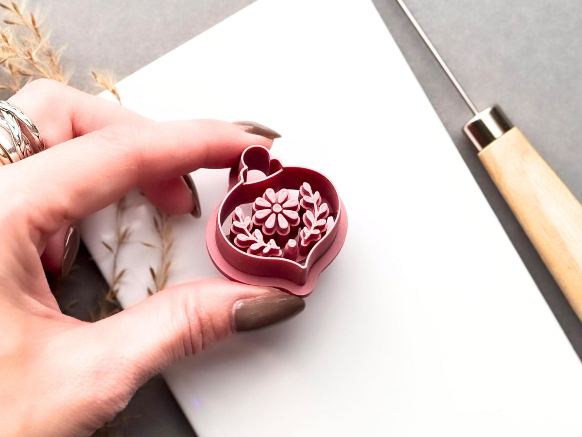 Floral Christmas bauble clay cutter held in a hand, featuring a central flower, leafy botanical stamp details and a hanging loop. Pink 3D-printed cutter shown close-up to highlight the raised cutting edge and intricate floral design.