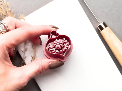 Floral Christmas bauble clay cutter held in a hand, featuring a central flower, leafy botanical stamp details and a hanging loop. Pink 3D-printed cutter shown close-up to highlight the raised cutting edge and intricate floral design.