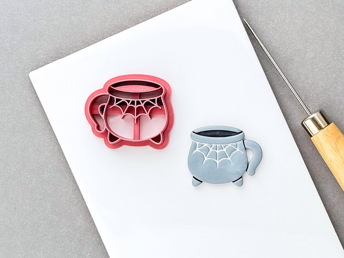 Halloween cauldron mug clay cutter featuring a bubbling witch’s cauldron with a spiderweb design, shown beside a finished grey polymer clay cauldron with matching web details and small feet. Spooky, witchy and Halloween-inspired design.
