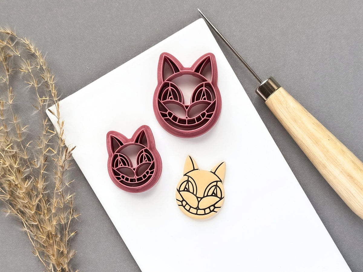 Stained glass style cat face polymer clay cutters displayed in two sizes, featuring pointed ears, stylised eyes, a rounded nose and segmented decorative details, shown beside a finished cream polymer clay cat face cut-out.