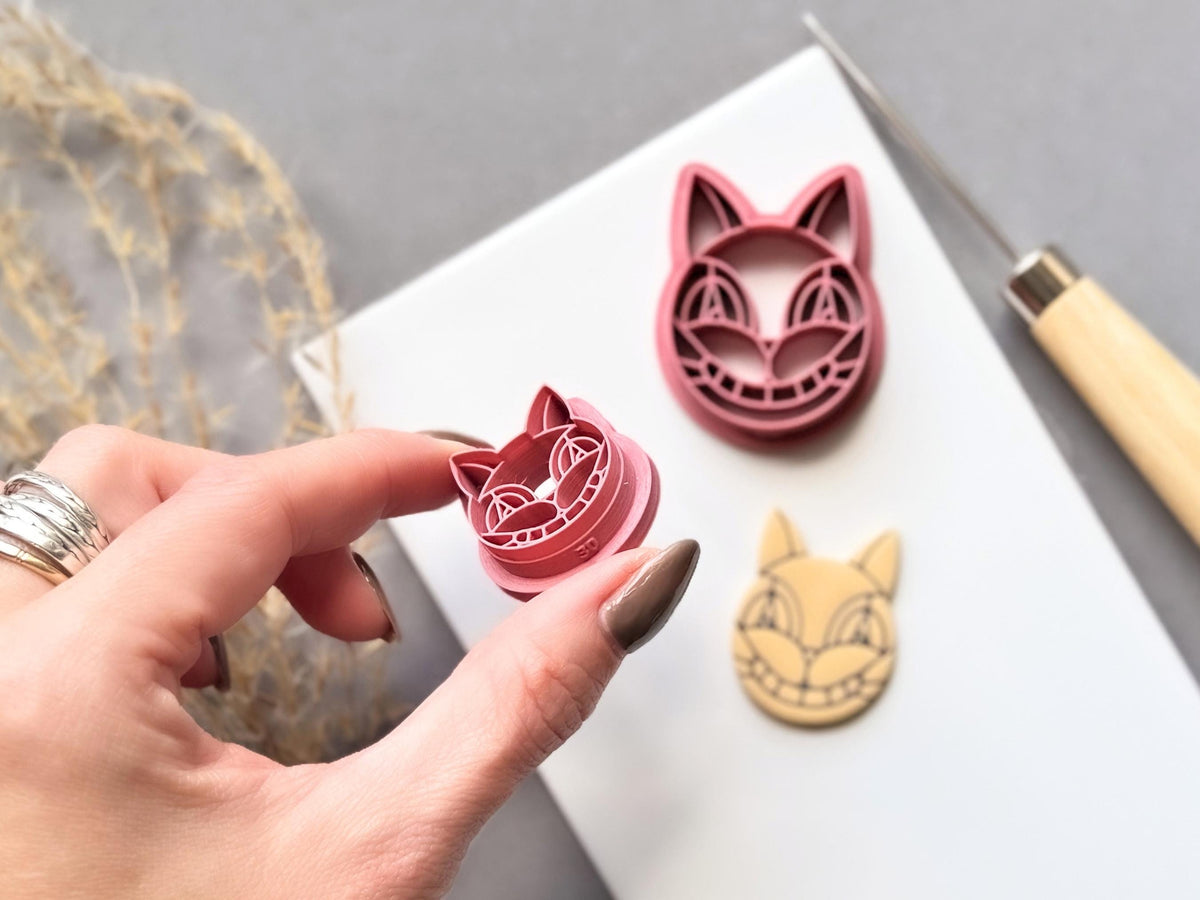 Stained glass style cat face clay cutter held in a hand, showing the detailed segmented design and raised cutting edge, with a larger cutter and finished cream polymer clay cat face cut-out in the background.